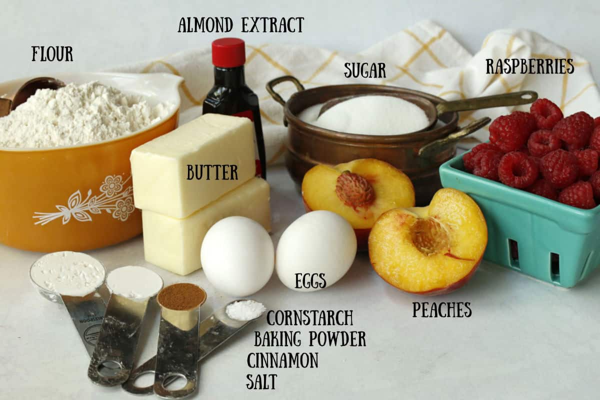 Baking ingredients including flour, butter, sugar, eggs, almond extract, cornstarch, baking powder, cinnamon, salt, fresh peaches, and raspberries are arranged on a counter.