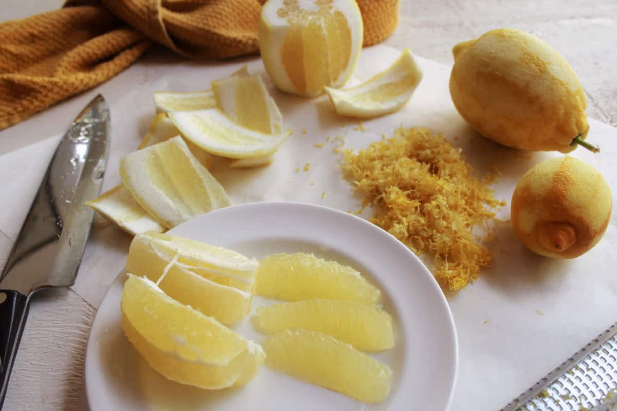 A peeled lemon, lemon segments on a plate, zest, knife, and whole lemons are arranged on a white surface with a brown cloth in the background.