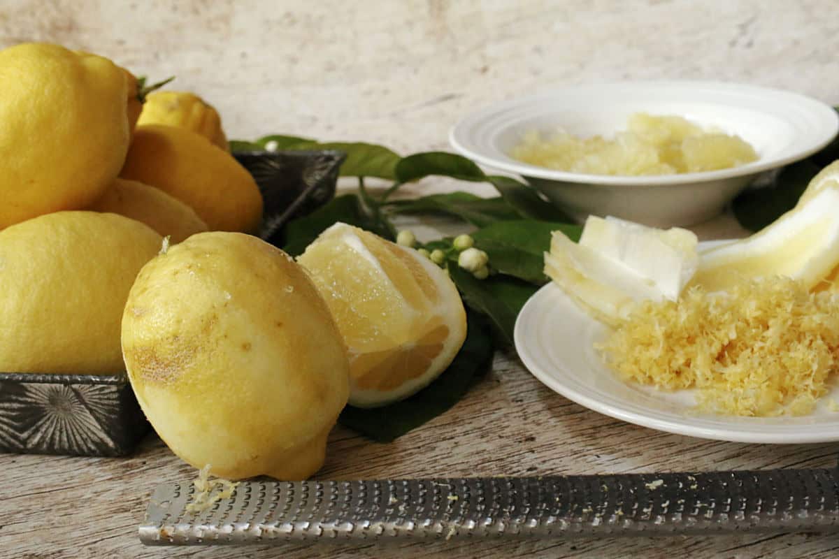 Whole and cut lemons, lemon zest, a grater, and lemon peel are arranged on a table with green leaves in the background.
