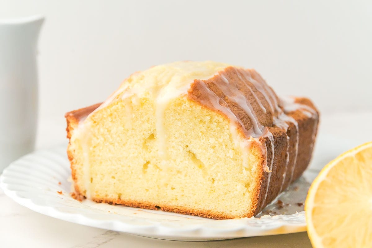 A sliced loaf of lemon pound cake with icing on top sits on a white plate next to a halved lemon.