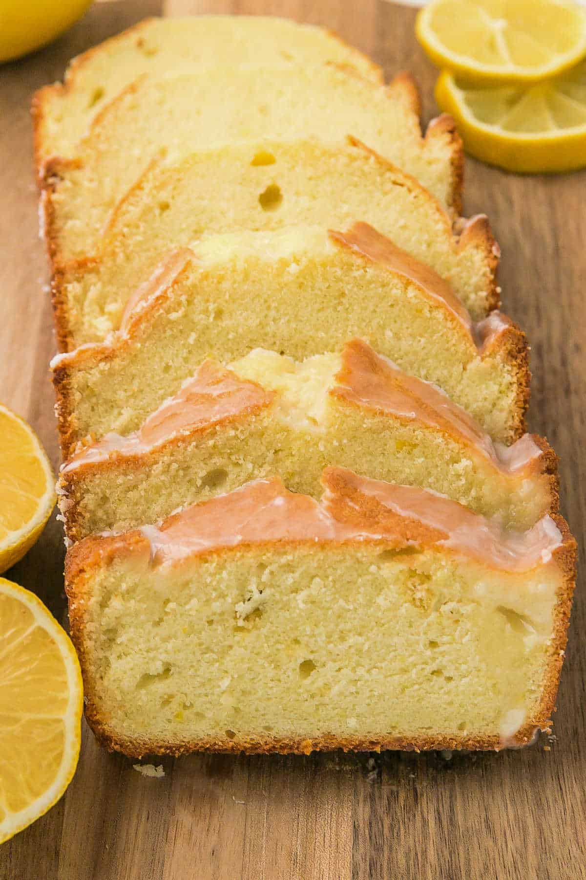 Sliced lemon loaf cake with a light glaze on a wooden surface, surrounded by halved lemons.