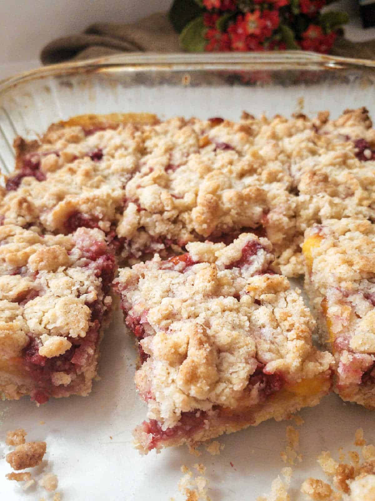 A glass baking dish contains several crumbly fruit bars with a streusel topping, and one bar has been removed from the dish.