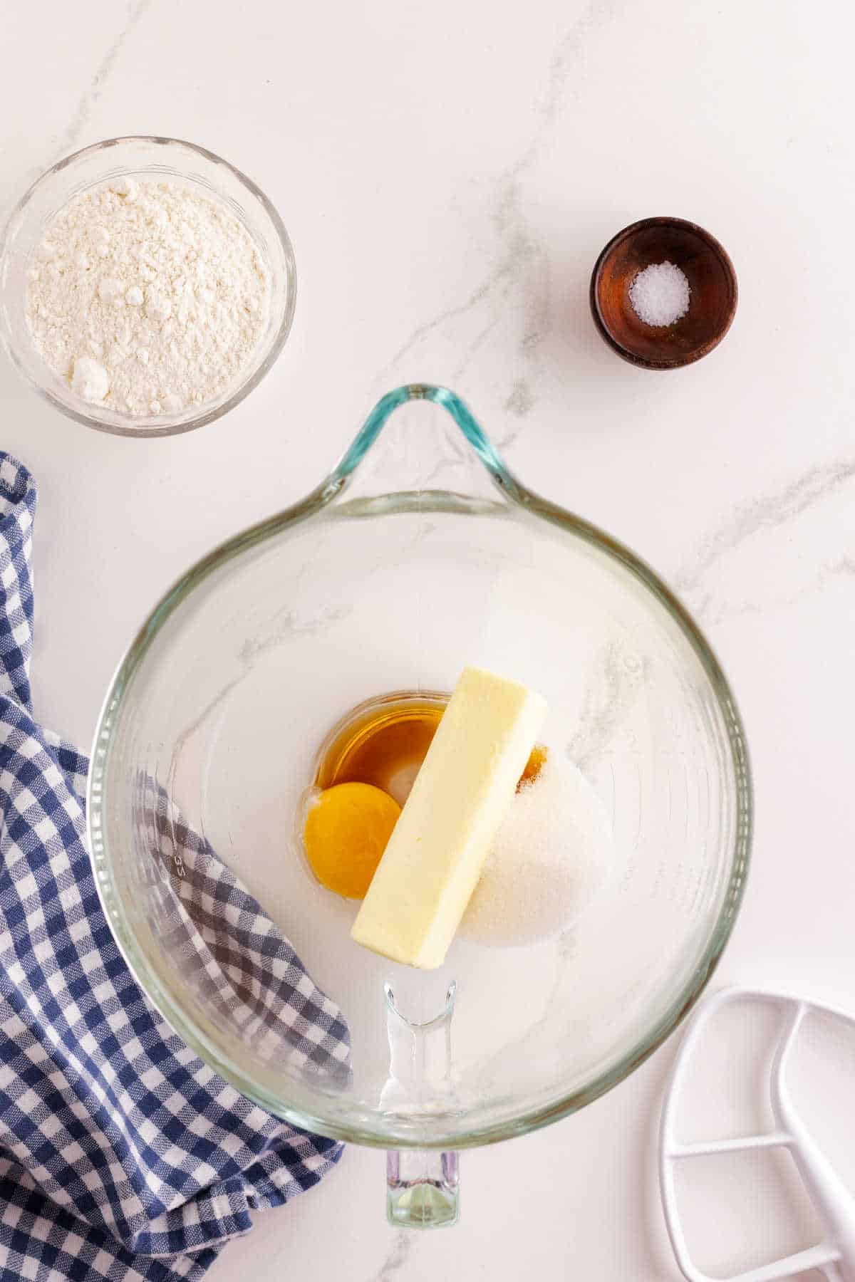 Top view of a glass mixing bowl with butter, sugar, egg, and vanilla next to a bowl of flour, a small bowl of salt, a mixer attachment, and a blue checkered cloth on a white surface.