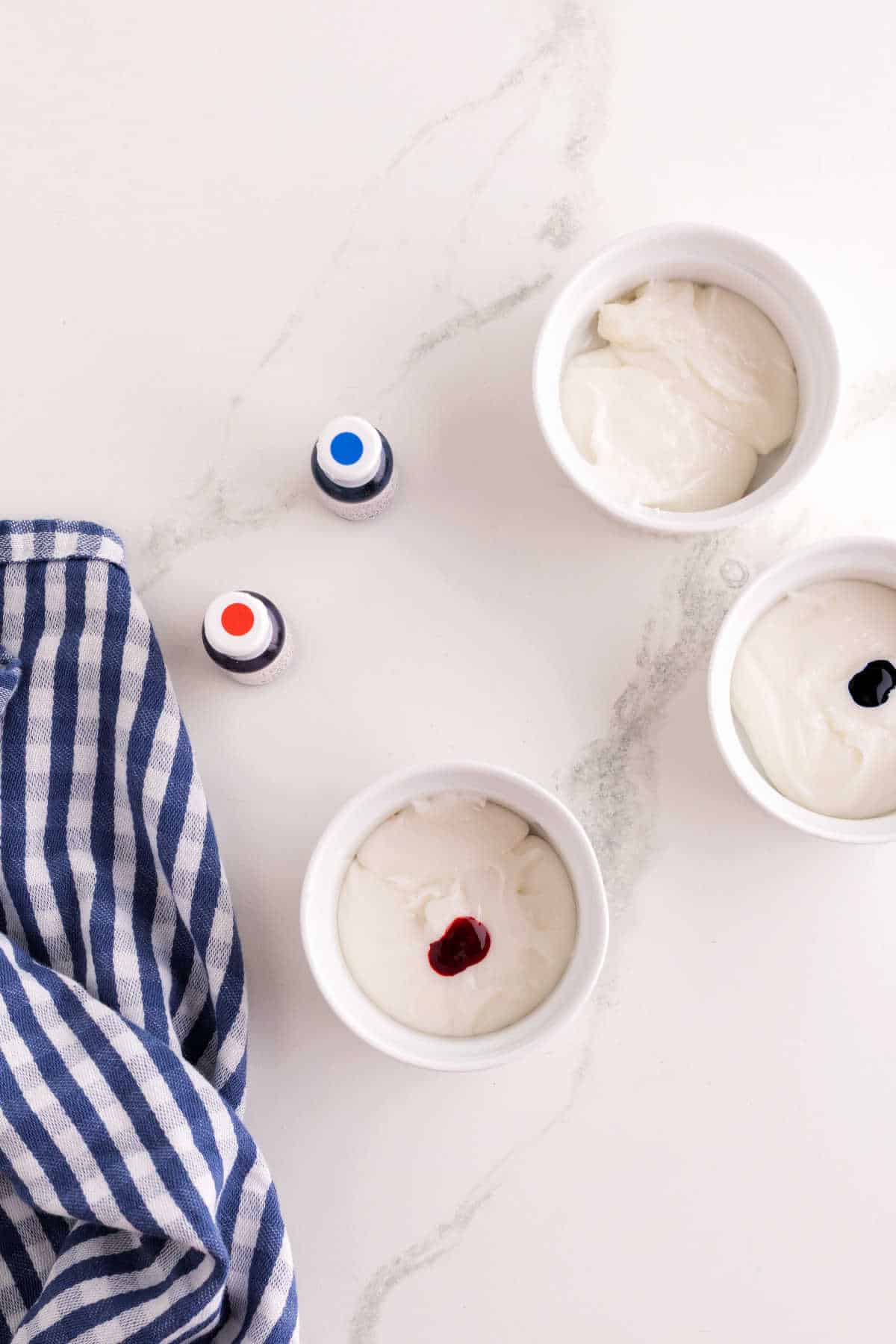 Three small bowls of white icing, two with drops of food coloring, sit on a marble surface next to blue and white striped cloth and two bottles of food coloring.