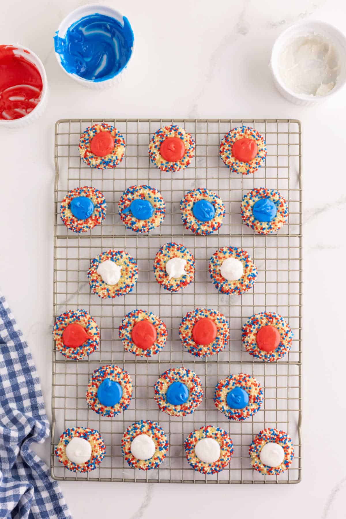 Cookies with red, white, and blue icing and rainbow sprinkles are arranged on a cooling rack. Small bowls of icing and a blue checkered cloth are nearby.