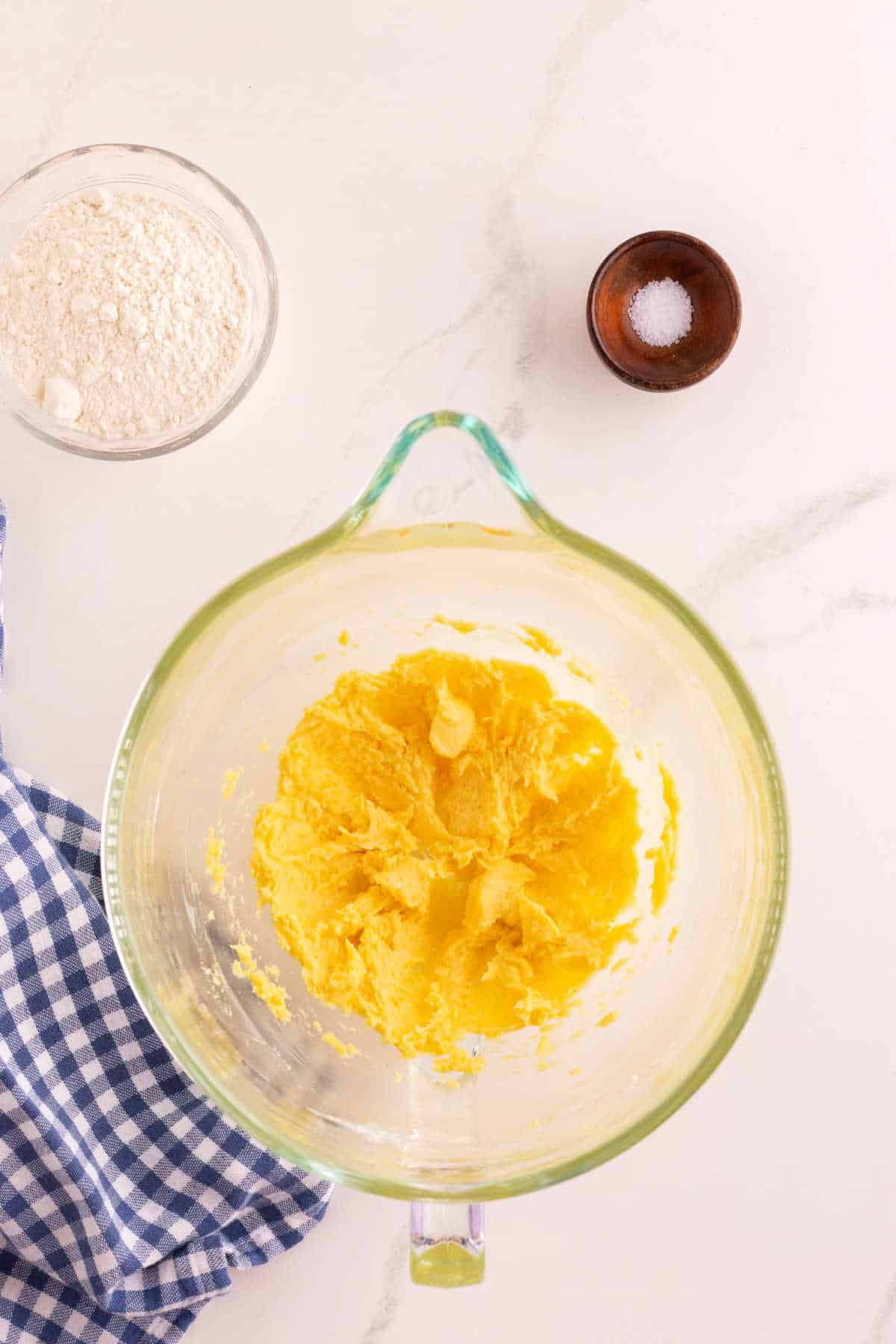 A glass mixing bowl with creamed butter and sugar, a small bowl of flour, a small dish of salt, and a blue checkered cloth on a white surface.