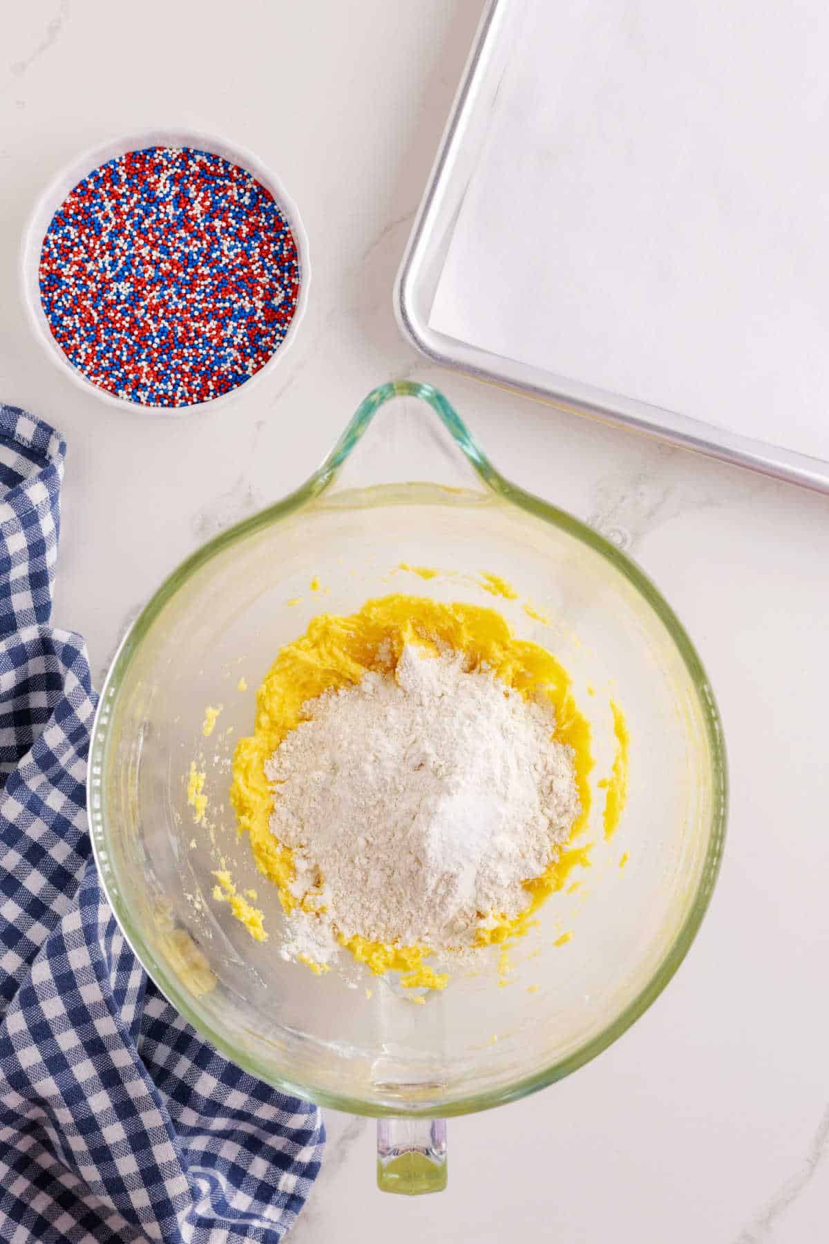 Glass mixing bowl with flour on top of yellow dough, next to a bowl of red, white, and blue sprinkles, a baking sheet, and a blue checkered towel on a white surface.