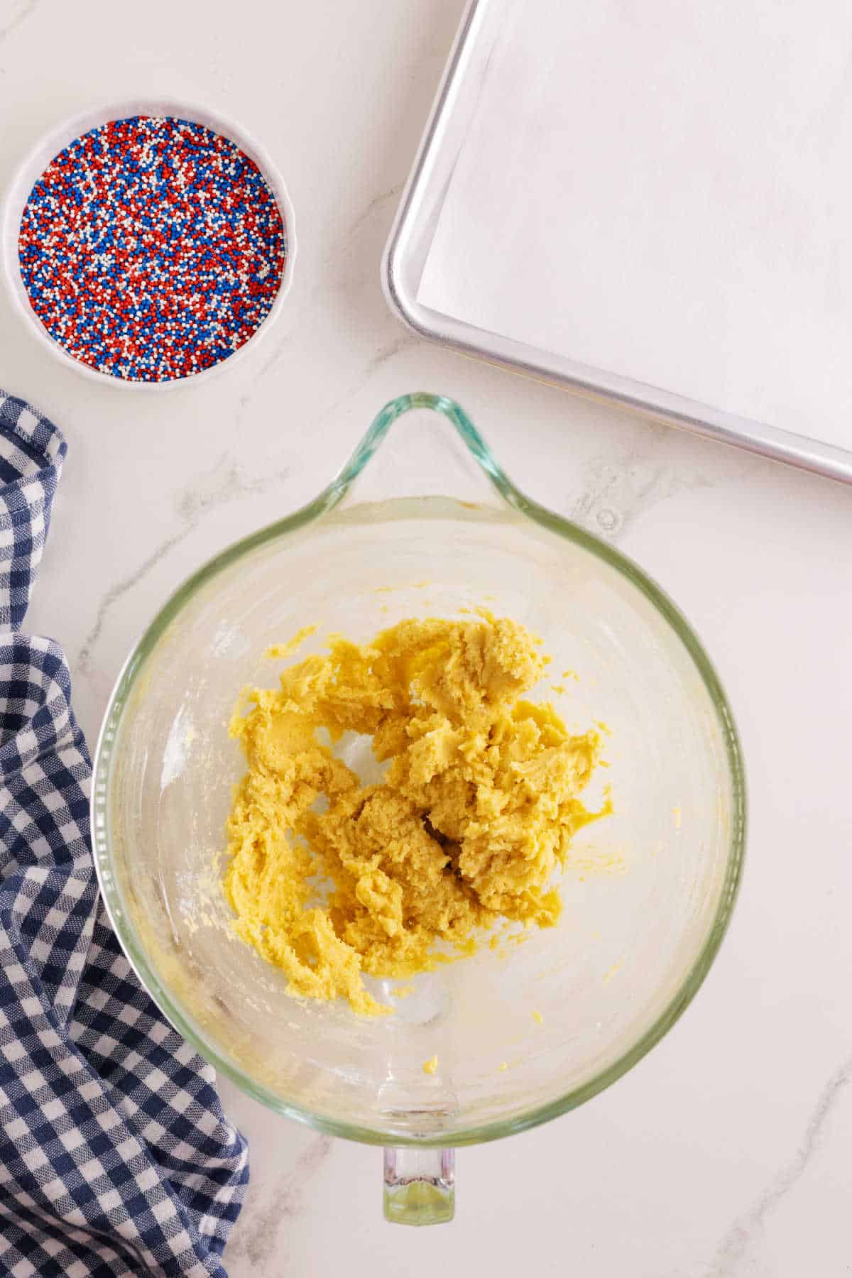 A glass mixing bowl with cookie dough, a bowl of red, white, and blue sprinkles, a baking sheet, and a checkered cloth on a marble countertop.