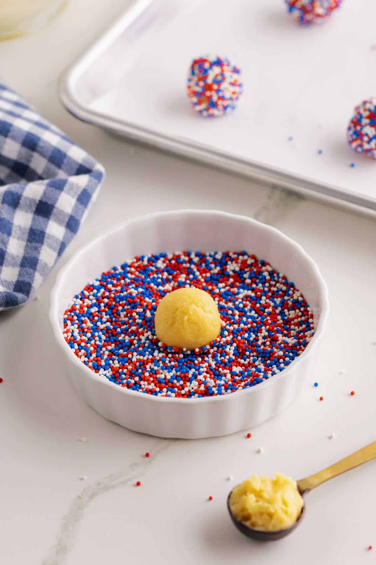 A ball of dough sits in a dish of red, white, and blue sprinkles. A spoon with dough and a baking tray with sprinkled balls are nearby on a white surface.