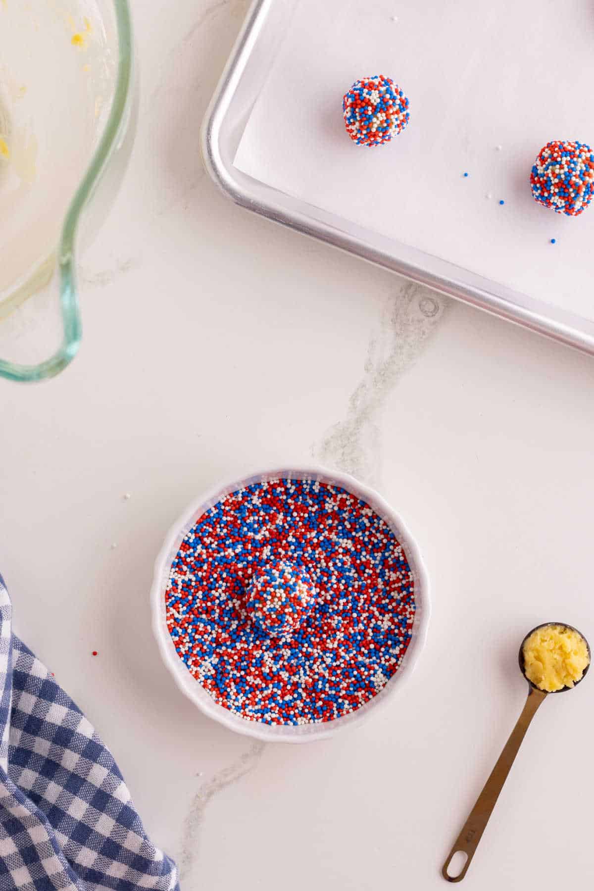 A bowl of red, white, and blue sprinkles next to a teaspoon of yellow batter, a baking sheet with sprinkled dough balls, and a blue checkered cloth on a white countertop.