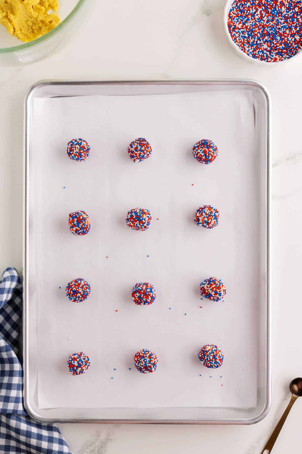 Twelve unbaked cookie dough balls coated with red, white, and blue sprinkles are arranged in rows on a parchment-lined baking sheet.