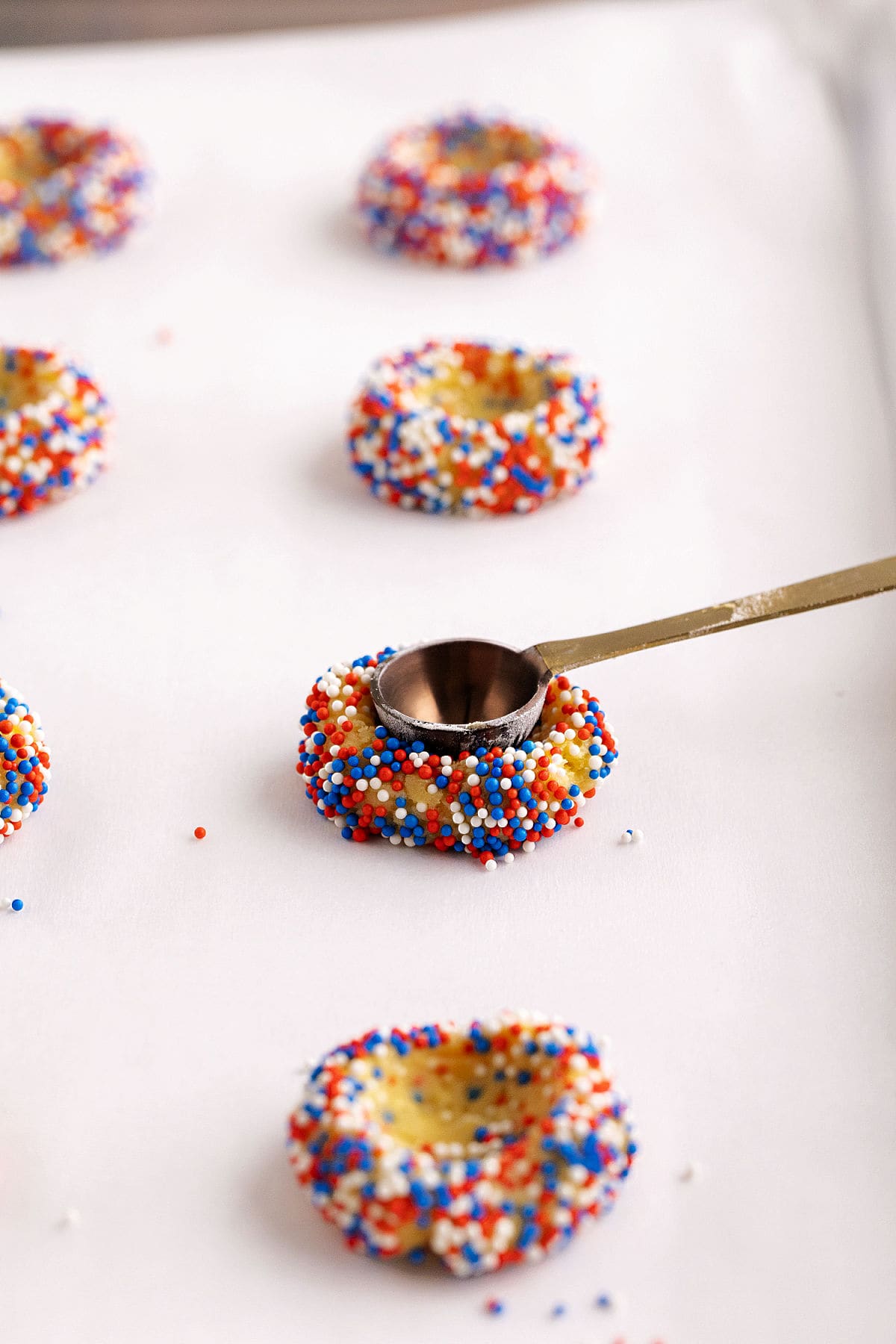 A metal spoon presses an indent into thumbprint sprinkle-covered cookie dough balls on a parchment-lined baking sheet.