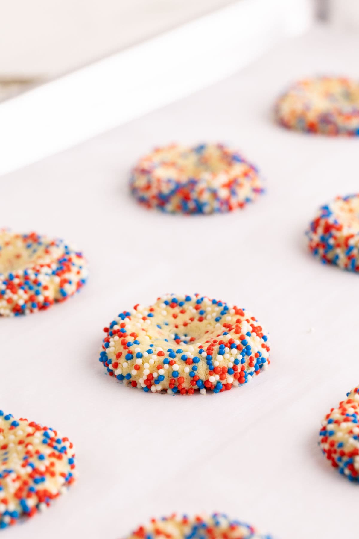 Baked thumbprint coated with red, white, and blue sprinkles are arranged on a parchment-lined baking sheet, ready to be baked.