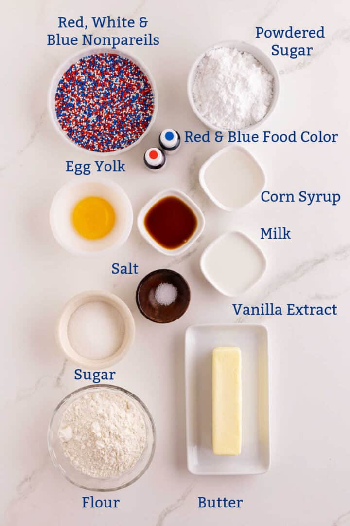 Assorted baking ingredients including nonpareils, powdered sugar, egg yolk, food coloring, corn syrup, milk, vanilla, salt, sugar, flour, and butter arranged on a white surface for thumbprint cookies.