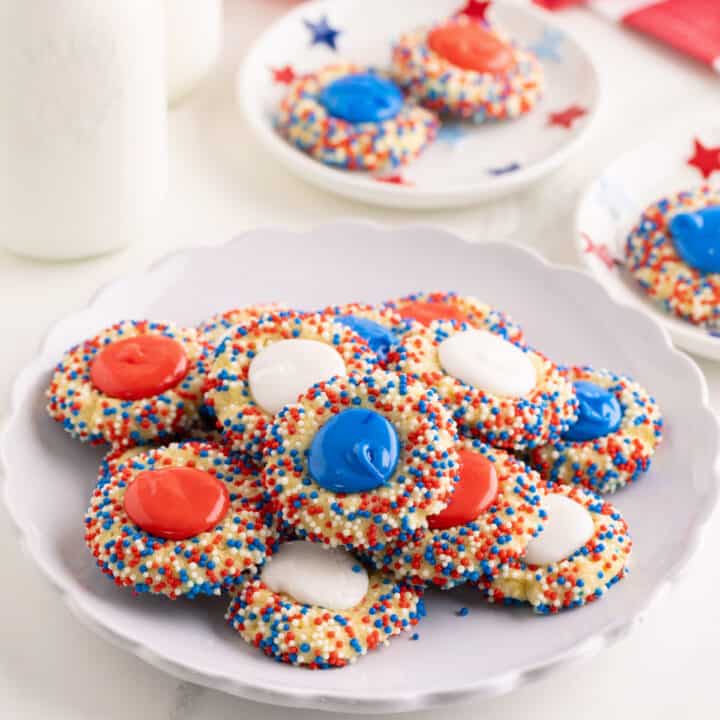 A plate of red, white and blue thumbprint cookies topped with red, white, and blue candy melts, and coated in matching sprinkles, sits on a white table with milk and more cookies in the background.