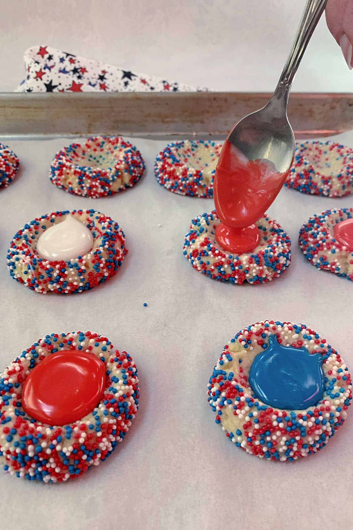 A hand uses a spoon to add red icing to thumbpint cookies coated in red, white, and blue sprinkles on a parchment-lined baking sheet.