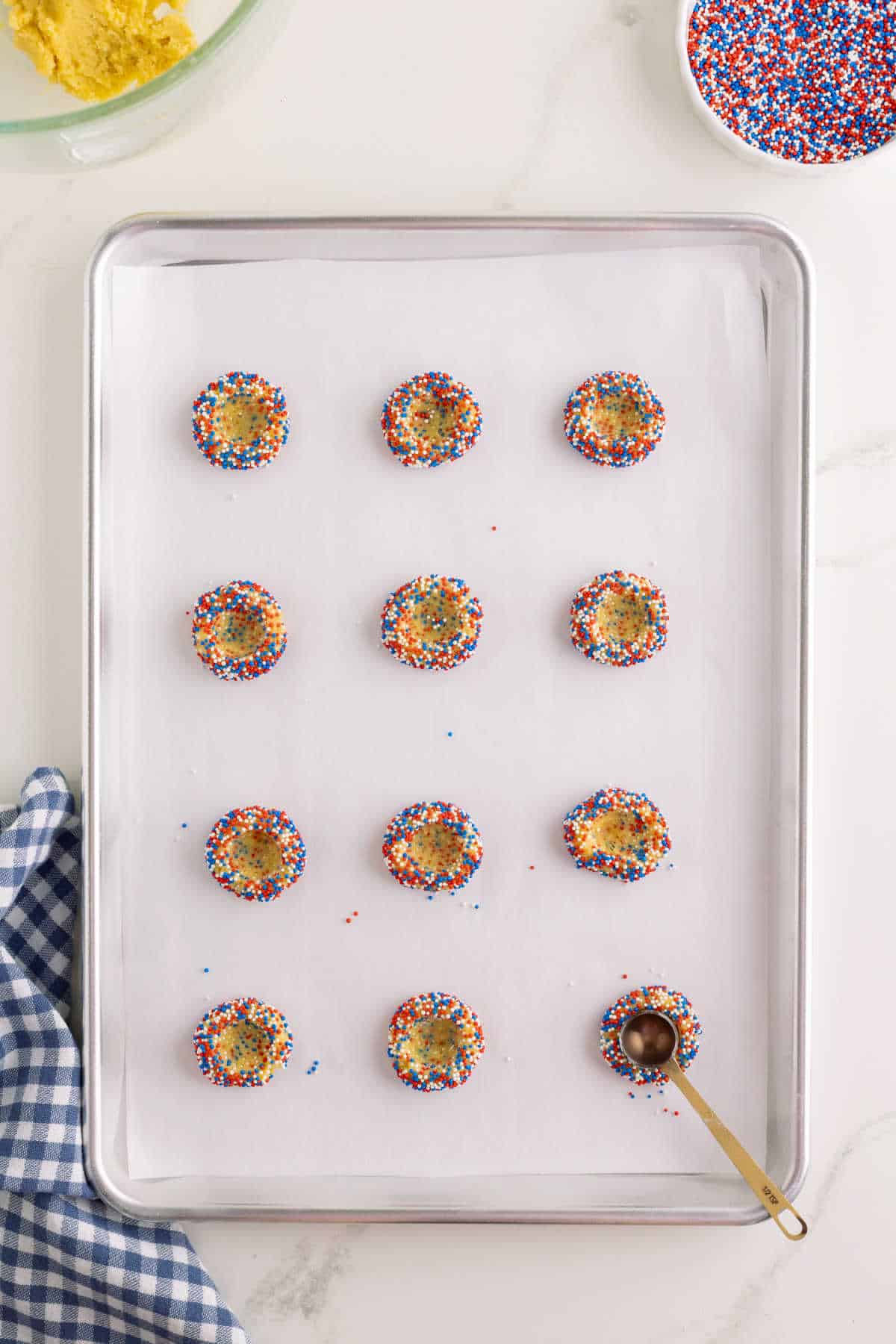 Twelve unbaked cookies coated with red, white, and blue sprinkles are arranged on a parchment-lined baking sheet, with a small measuring spoon beside them.