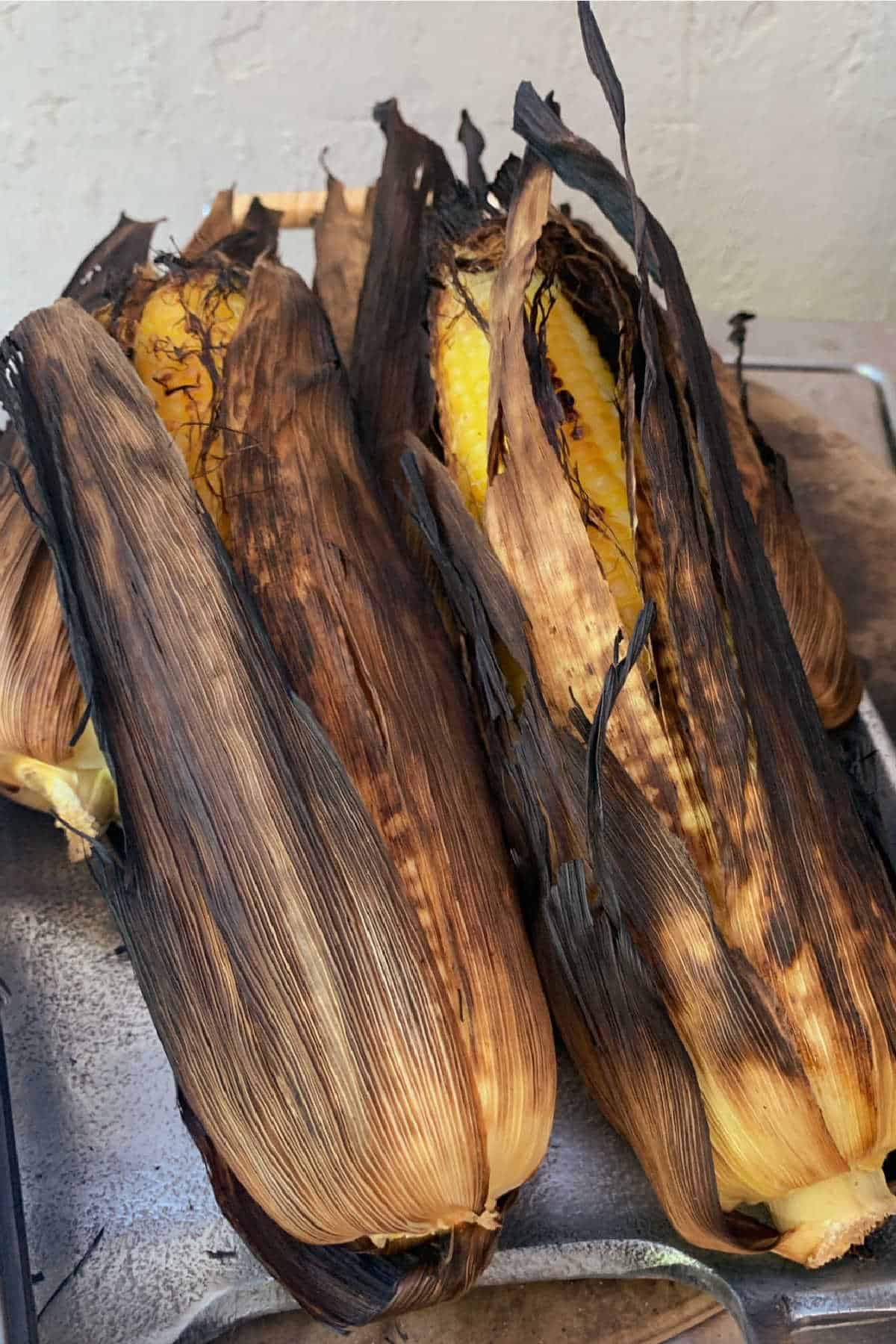 Three ears of corn with partially charred husks, grilled and resting on a metal surface. The yellow kernels are visible through the open husks.