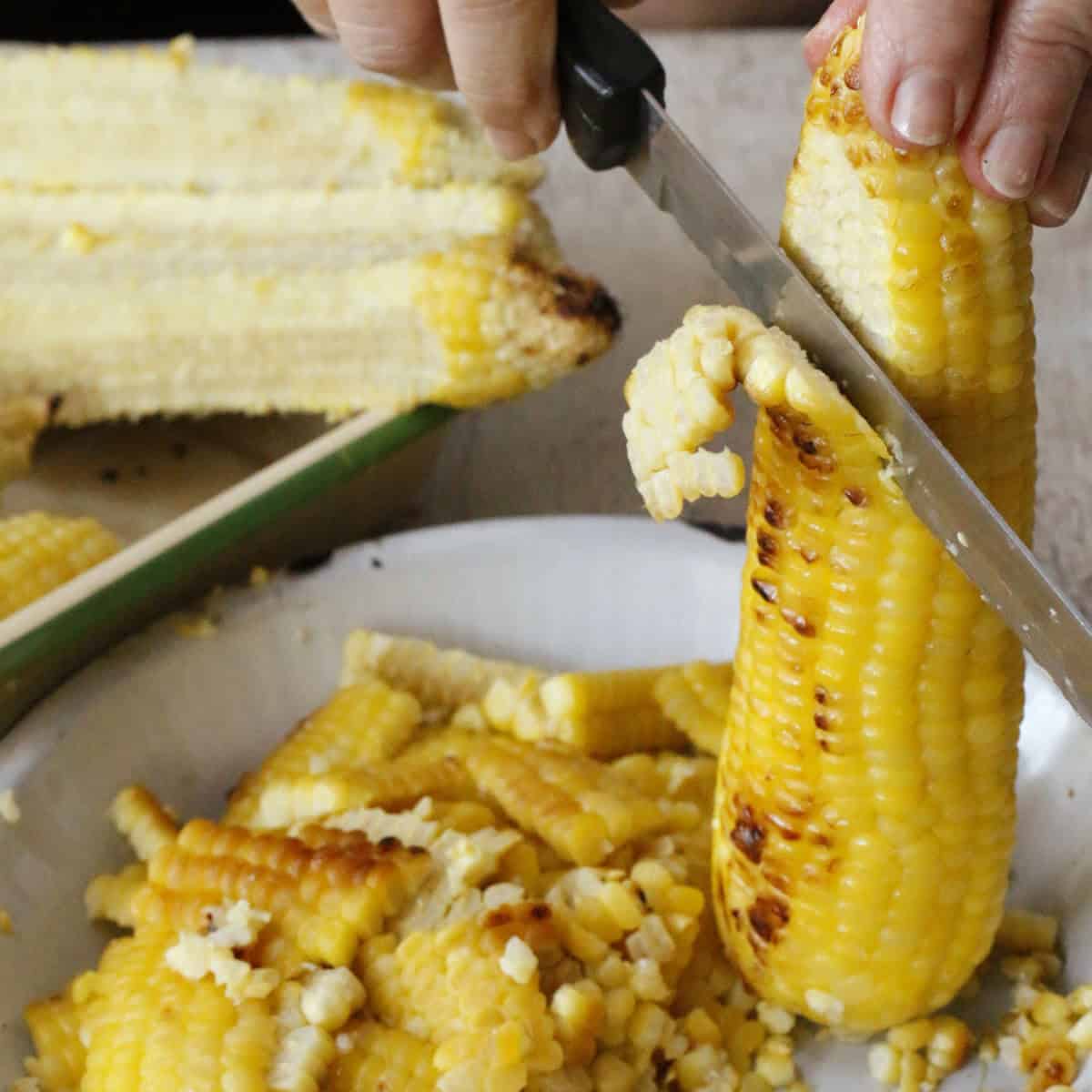 A person cuts kernels off a cooked corn cob with a knife over a plate filled with corn kernels.