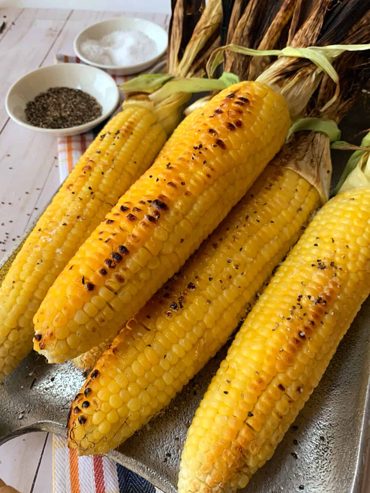 Grilled corn on the cob with char marks, arranged on a tray with bowls of salt and pepper in the background.