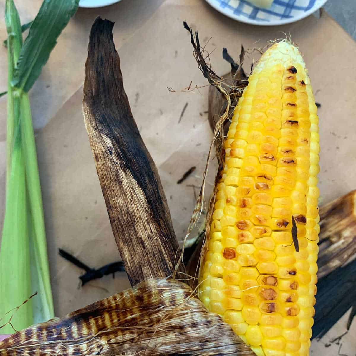 A grilled ear of corn with partially charred husk, placed on brown paper, with a plate in the background.