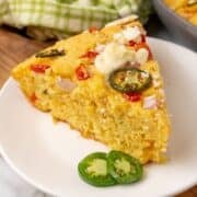 A slice of cornbread topped with cheese, sliced jalapeños, onions, and tomatoes is served on a white plate with extra jalapeño slices.