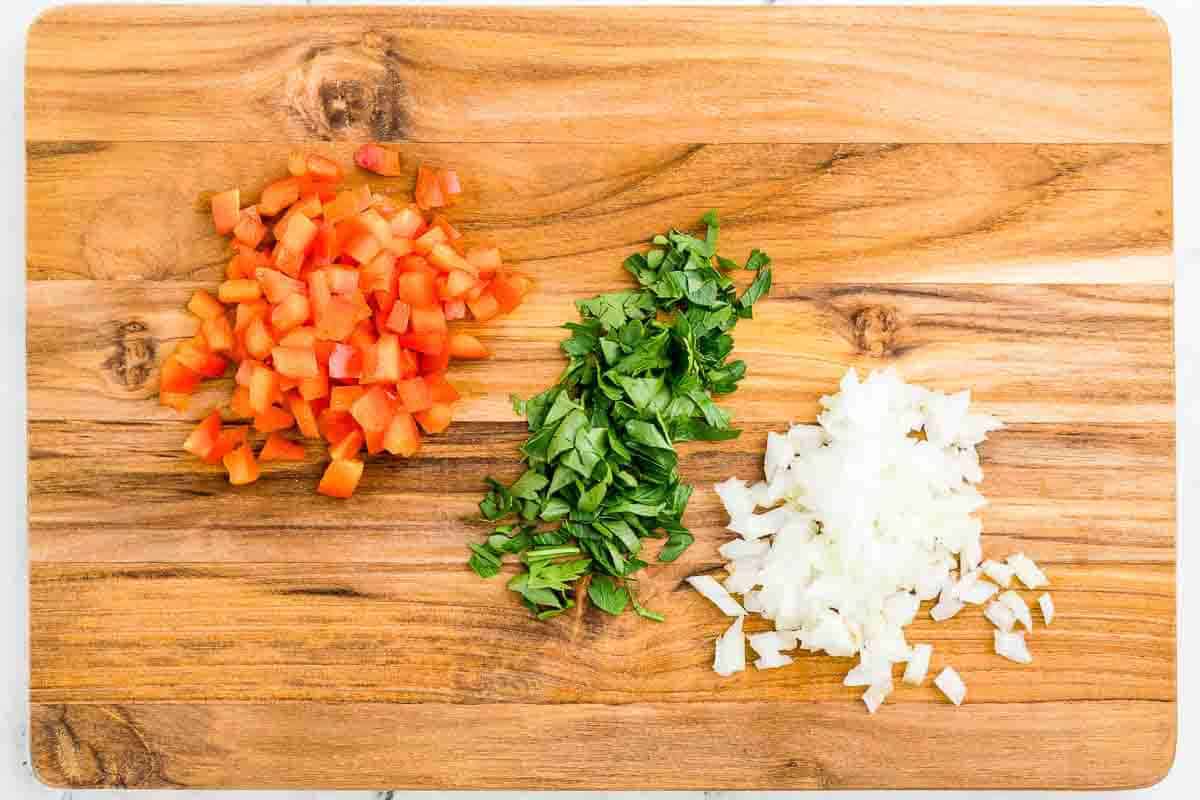 Chopped tomatoes, fresh basil, and diced onions arranged in separate piles on a wooden cutting board.
