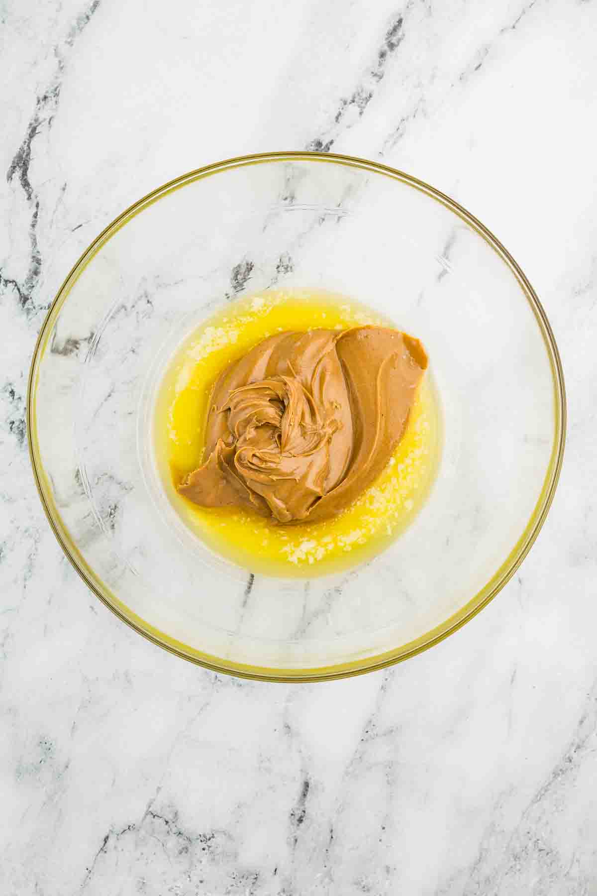 A glass bowl on a marble surface contains melted butter and creamy peanut butter.