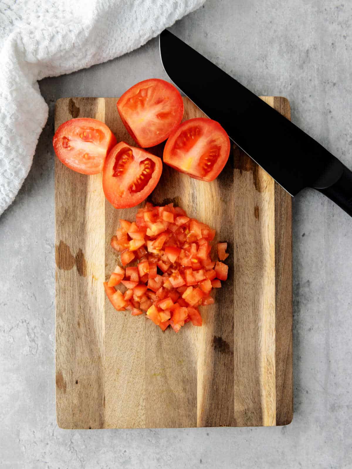 A wooden cutting board with diced tomatoes, halved tomatoes, a black knife, and a white cloth on a gray countertop.