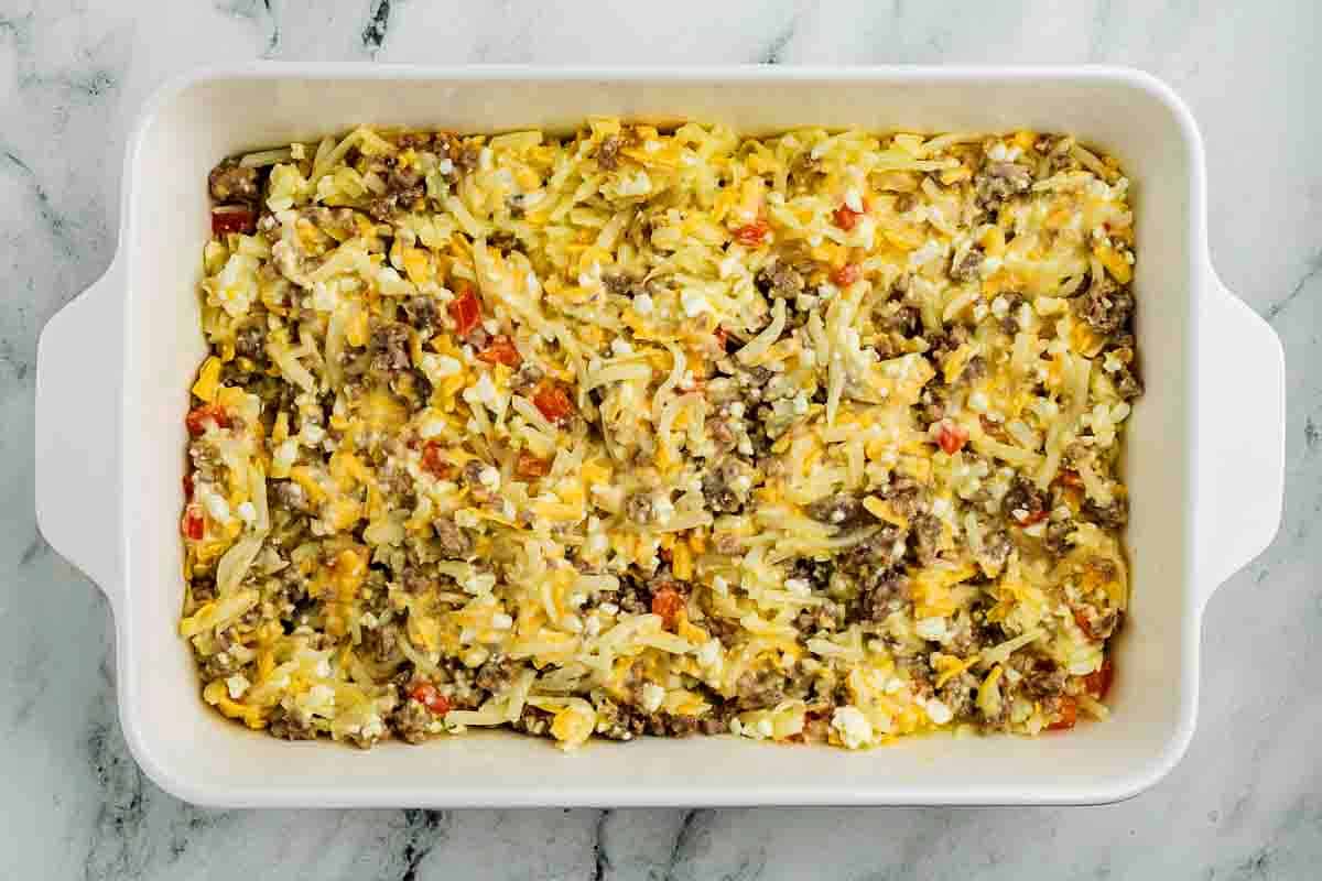 A white baking dish filled with a baked casserole containing shredded potatoes, ground meat, melted cheese, and diced red peppers on a marble surface.
