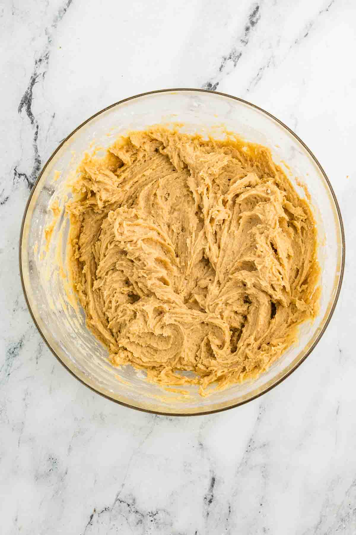 A glass bowl filled with light brown cookie dough sits on a white marble surface.