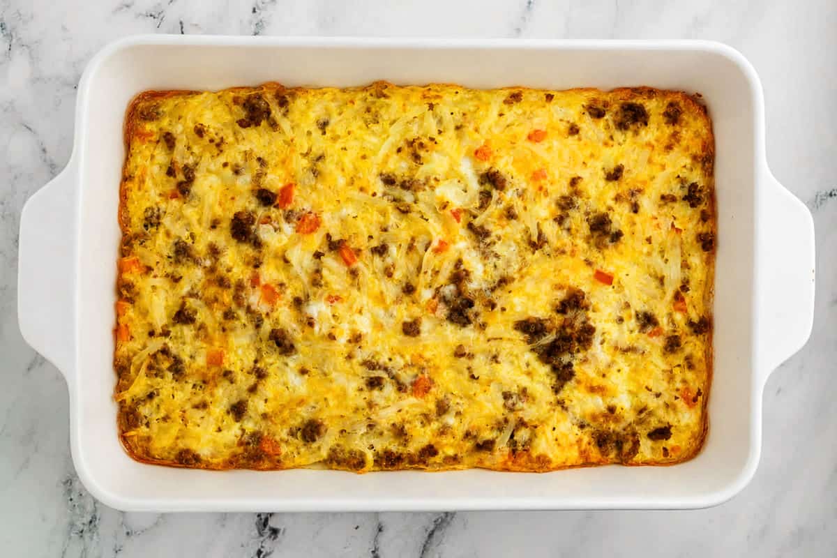 A baked breakfast casserole in a white rectangular dish, containing eggs, sausage, cheese, and diced vegetables, on a marble surface.