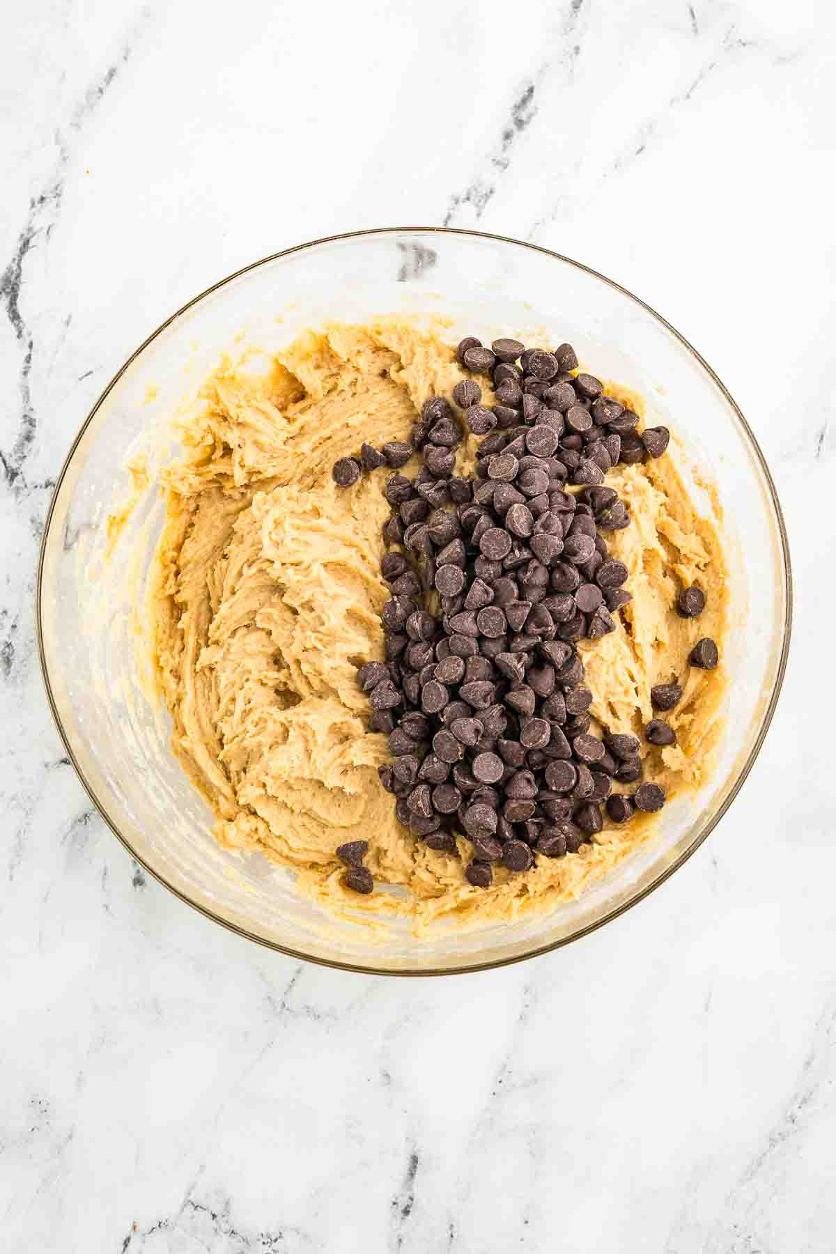 A glass bowl with cookie dough and a pile of chocolate chips on top sits on a white marble surface.