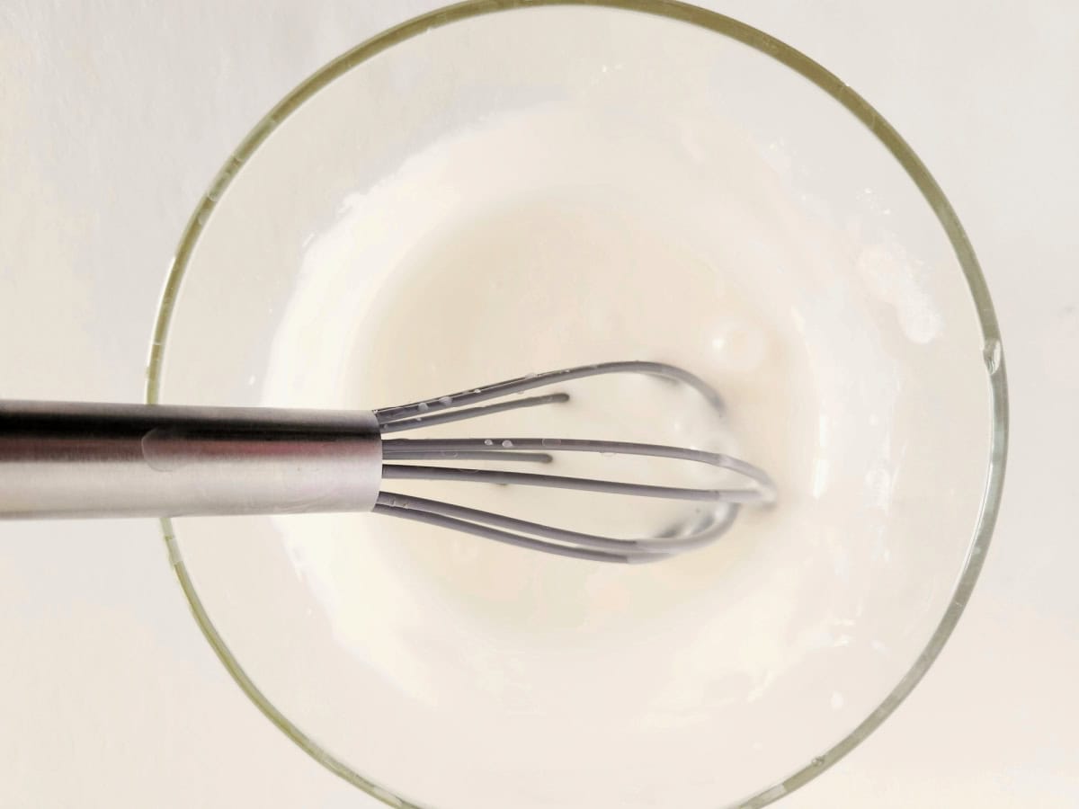 A metal whisk is mixing a white liquid, possibly batter or cream, in a clear glass bowl viewed from above against a light background.