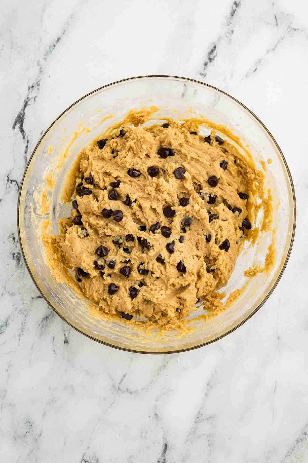 A glass bowl filled with chocolate chip cookie dough sits on a white marble surface.