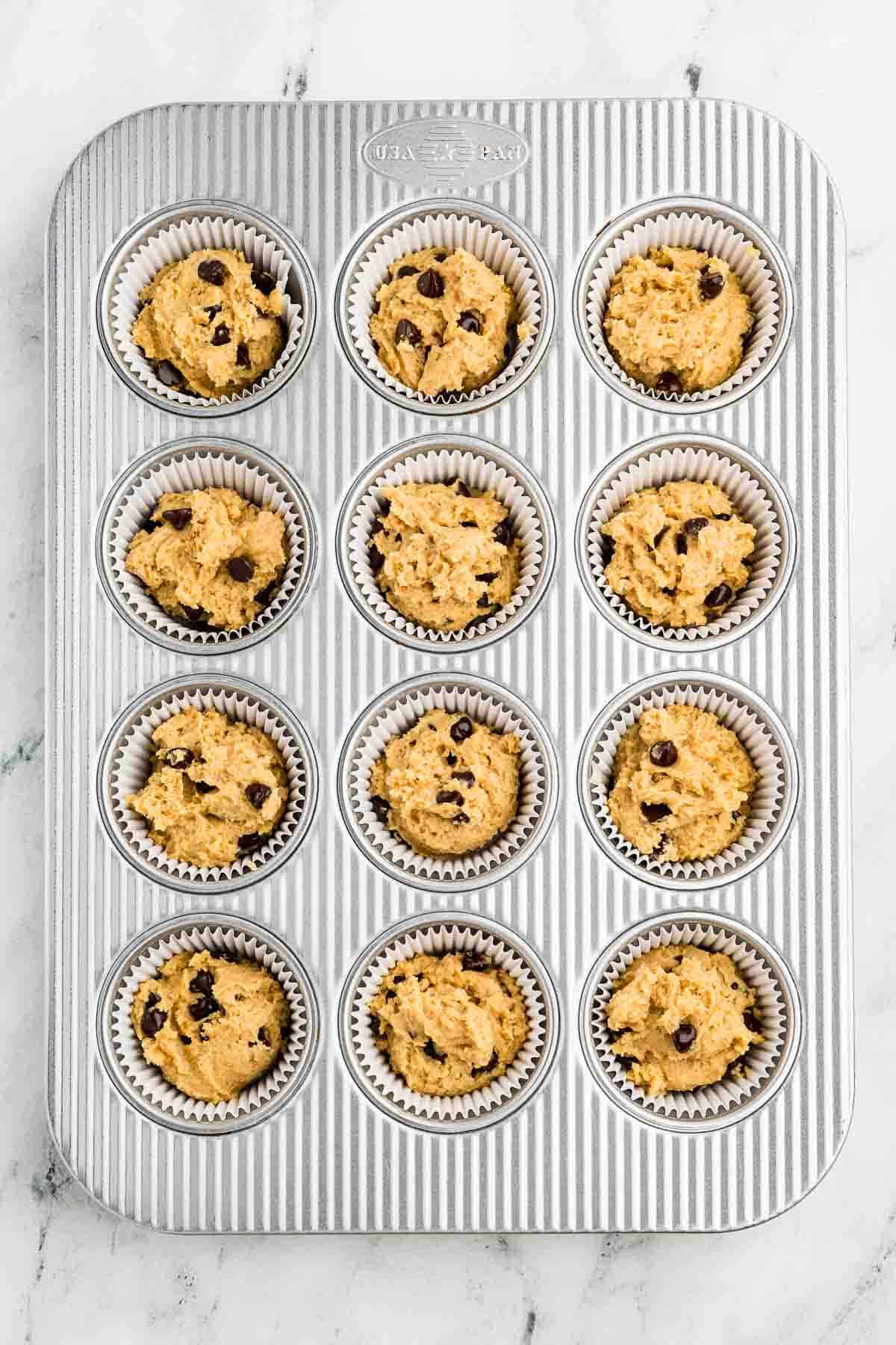 A muffin tin with twelve paper liners, each filled with unbaked chocolate chip muffin batter, is placed on a marble surface.