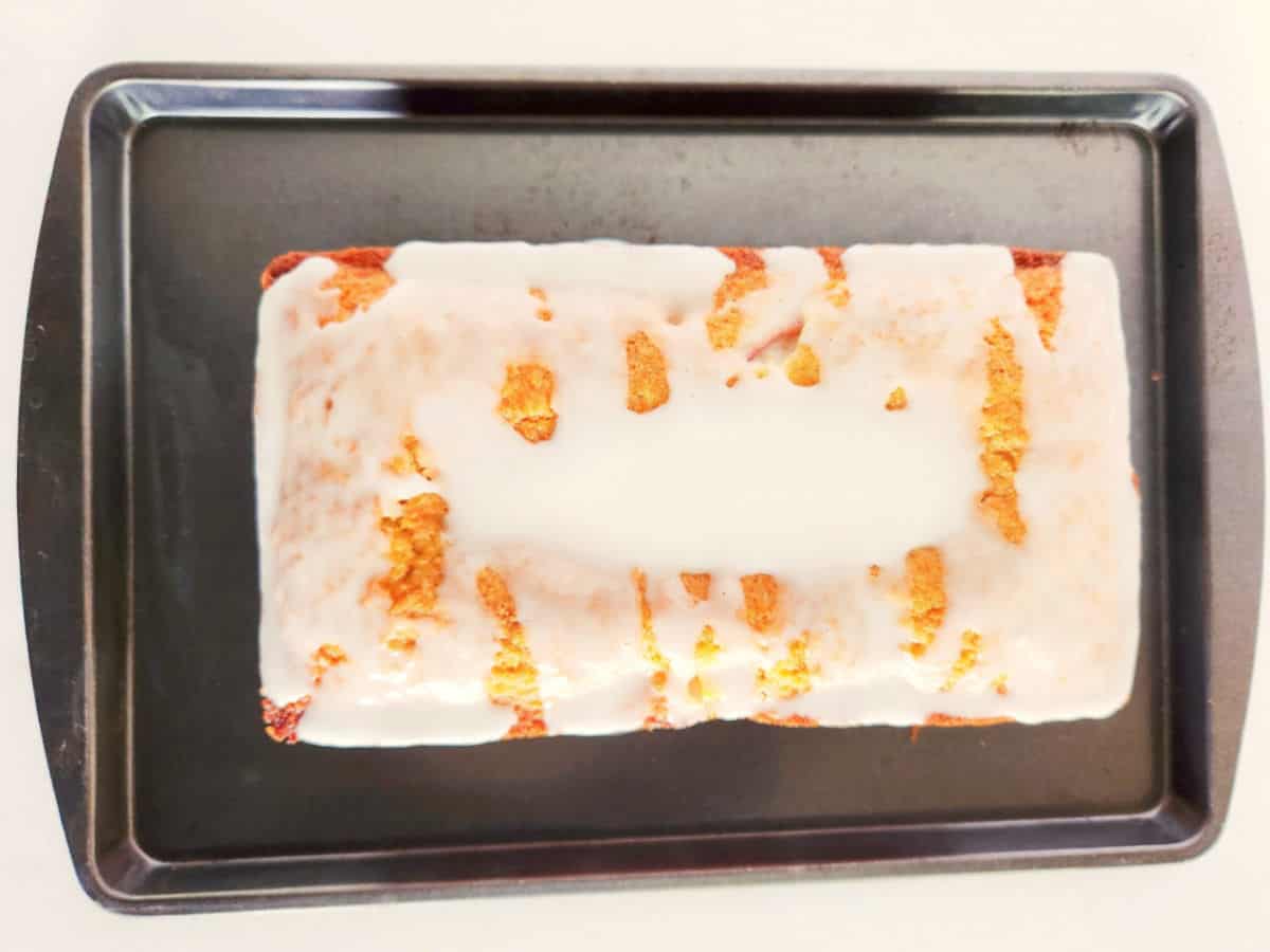 A rectangular peach loaf cake with white icing is placed on a black baking tray. The icing covers the top unevenly, revealing parts of the golden-brown cake underneath.