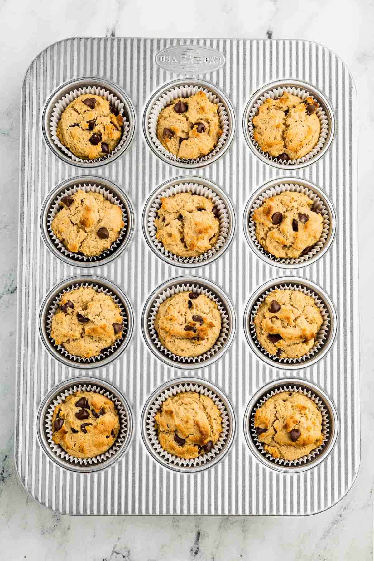 A metal muffin tray holds 12 paper liners filled with baked peanut butter chocolate chip muffins, some with visible chocolate chips, on a light marble surface.