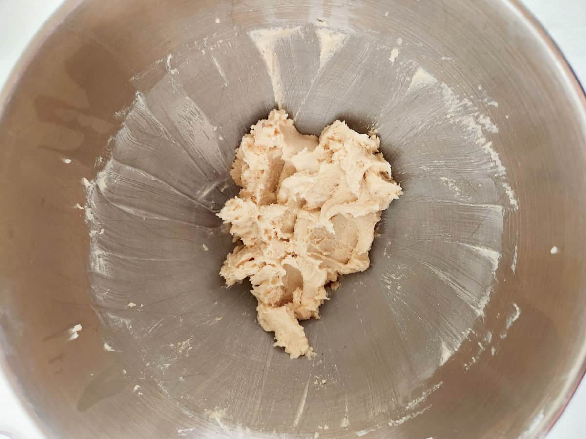 A mound of cookie dough sits in the bottom of a large, brushed metal mixing bowl, with remnants of dough spread along the sides.