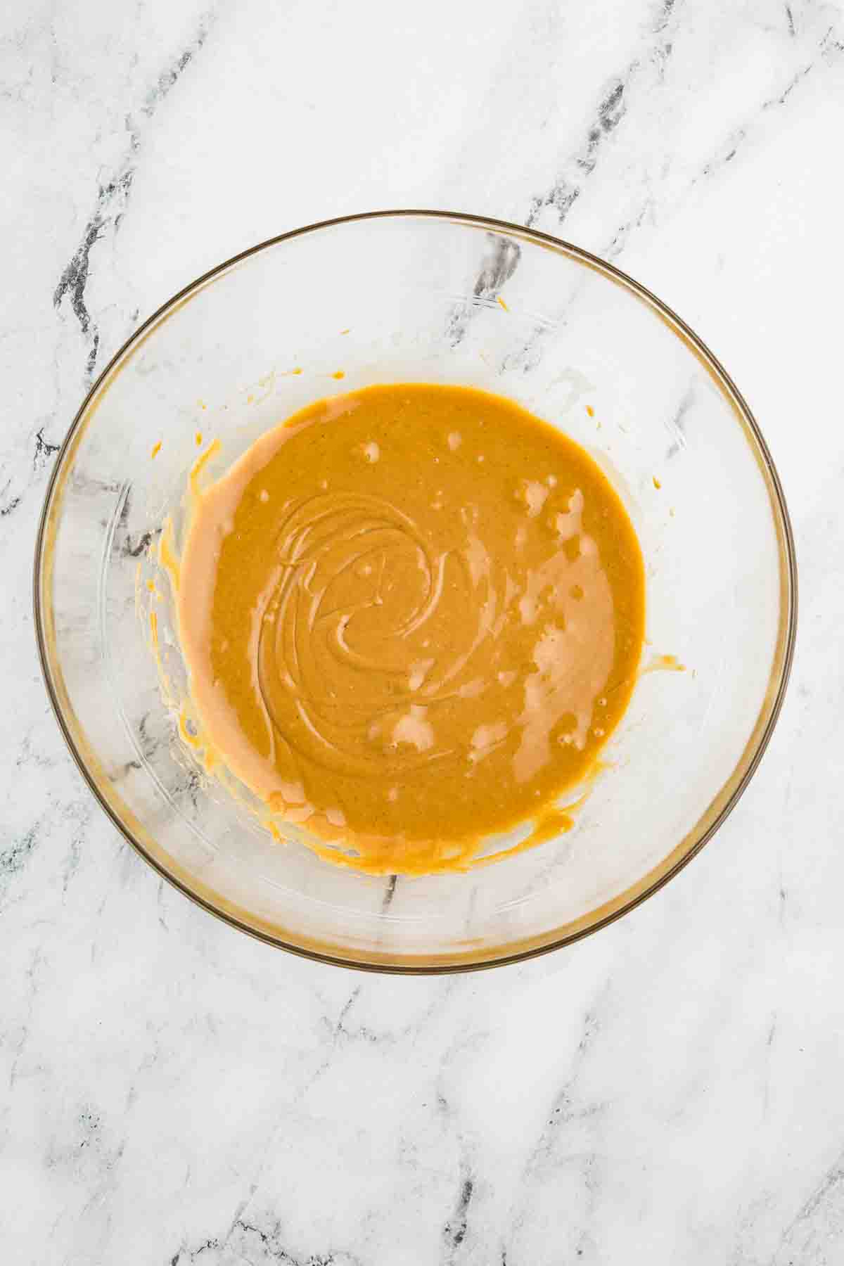 A glass bowl contains a light brown, creamy mixture with visible swirls, placed on a white marble surface.