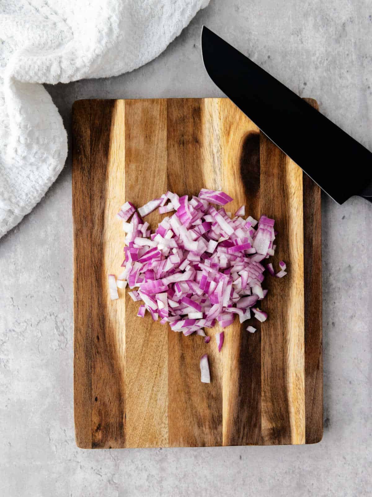 A pile of diced red onion on a wooden cutting board next to a black knife, with a white textured cloth nearby.