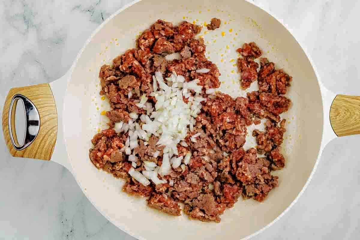 Ground beef and chopped onions cooking in a white skillet with wooden handles on a marble surface.