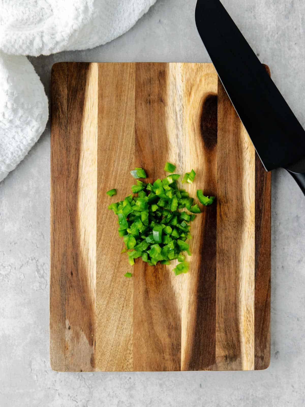 Chopped green bell pepper on a wooden cutting board, next to a black knife and a white towel on a gray countertop.