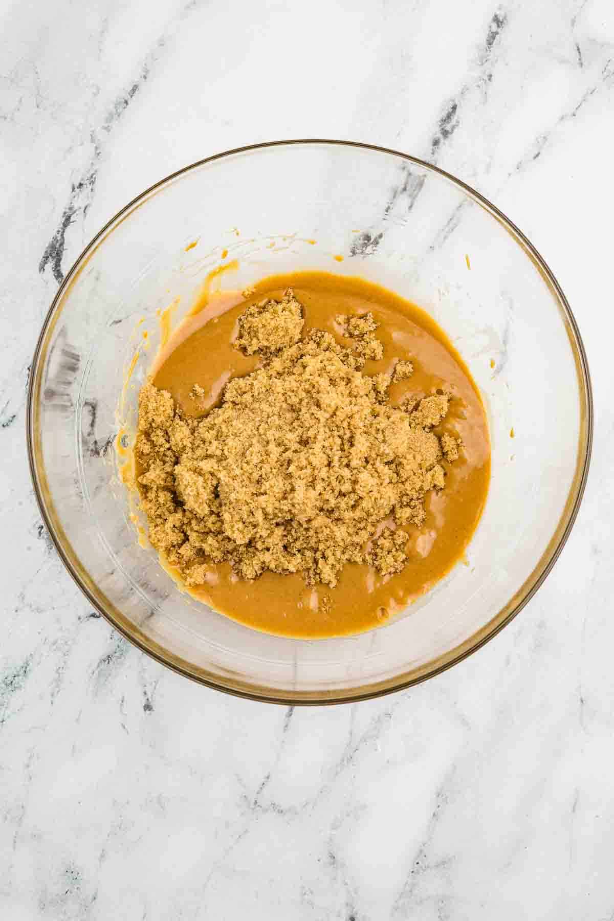 A glass bowl containing a mixture of brown sugar and a caramel-colored liquid sits on a white marble surface.