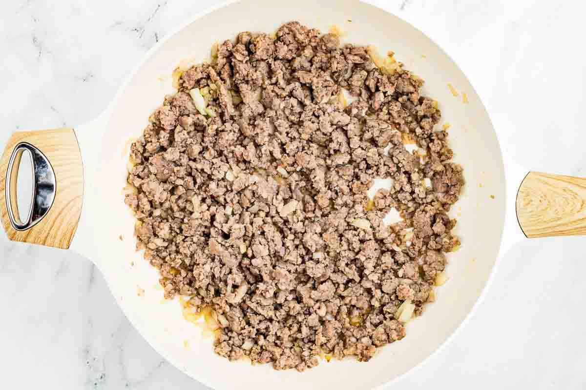 Cooked ground beef with diced onions in a white skillet, shown from above on a light countertop.