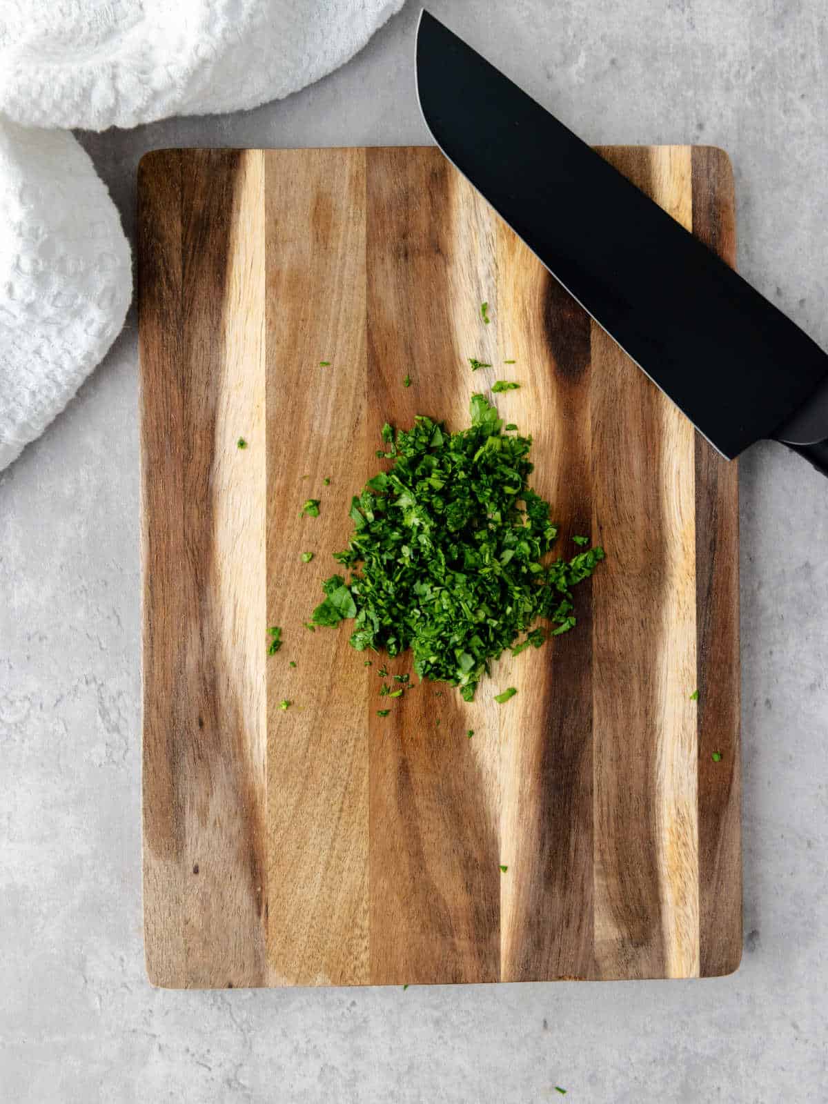 Chopped herbs on a wooden cutting board next to a black knife, with a white towel in the top left corner.