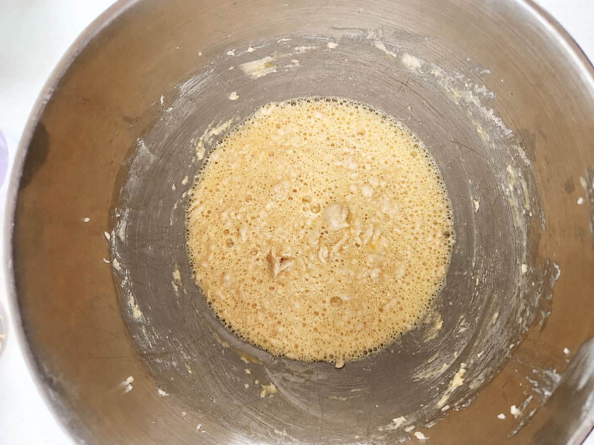 A metal mixing bowl containing a light brown, frothy liquid batter with some lumps and batter residue on the sides of the bowl.