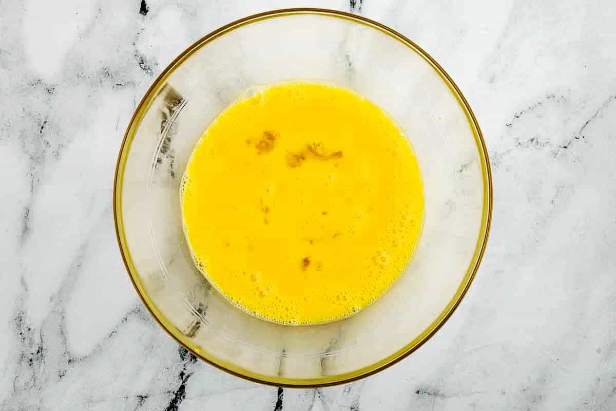 A glass bowl containing beaten eggs sits on a white marble surface.