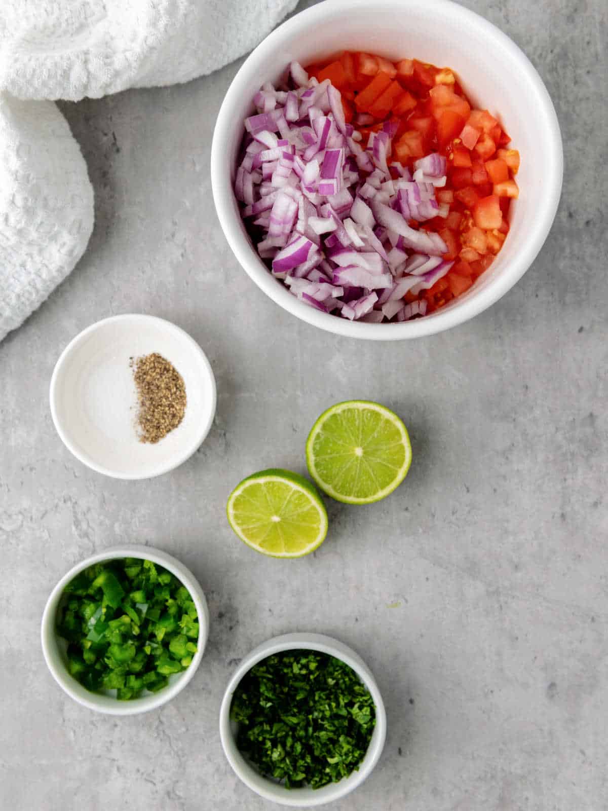 Chopped red onion, tomato, jalapeño, and cilantro in bowls, with halved limes and a small dish of ground pepper on a gray surface.