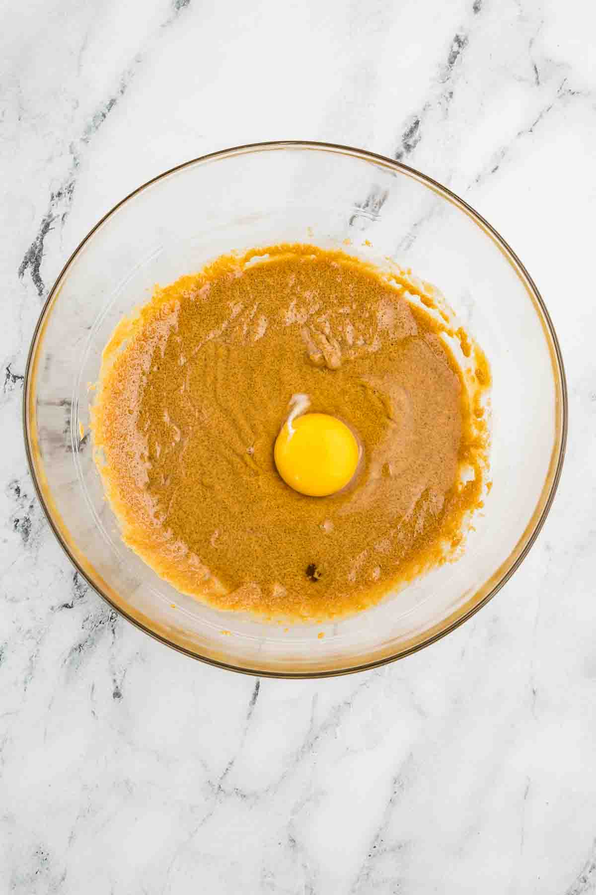 A glass bowl with brown sugar mixture and a raw egg in the center, placed on a white marble surface.
